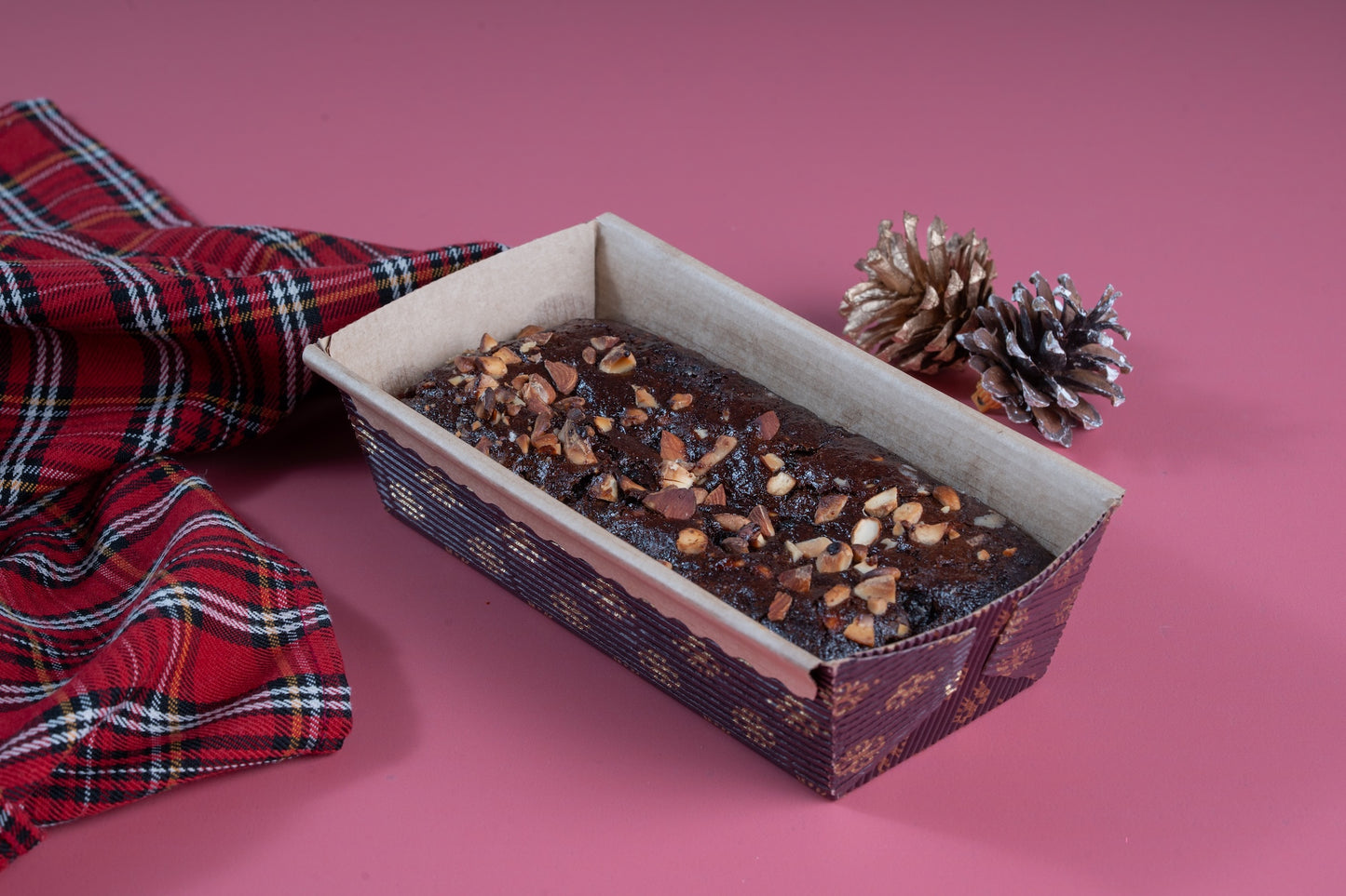 Whisk & Wish vegan rum raisin cake topped with chopped nuts, baked in a loaf tin and styled on a pink background with a red plaid cloth and pine cones, highlighting a premium plant-based dessert.