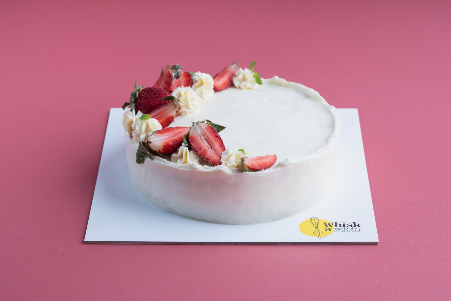 Whisk & Wish whole wheat sugar-free vanilla strawberry cake topped with fresh strawberries and cream, placed on a white cake board against a pink background, showcasing a healthy handcrafted dessert.